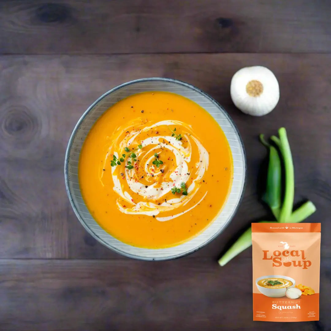 Bowl of orange soup with a swirl of cream, garlic, and green onions on a wooden surface, next to a 'Local Soup' package.
