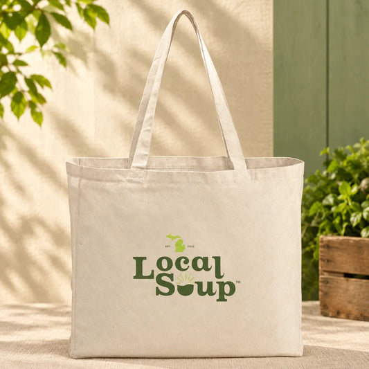 A natural canvas tote bag with the 'Local Soup' logo printed in green on the front, set against a rustic background with a wooden wall, green foliage, and a potted plant.